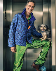 A forty years old man wears avant garde clothing. He has fun with his little dog in an modern elevator. AI generated.