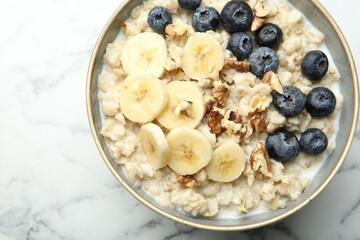 Tasty oatmeal with banana, blueberries, walnuts and milk served in bowl on white marble table, top view