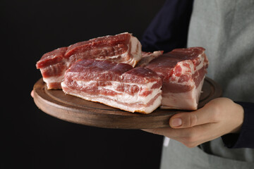 Woman holding wooden board with pieces of raw pork belly on black background, closeup