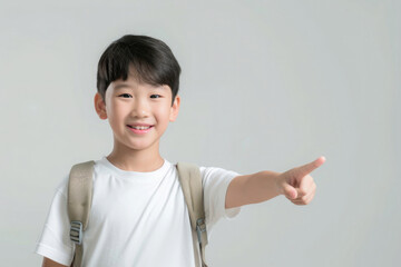 a boy with an Asian appearance in a white T-shirt with a briefcase, a schoolboy, smiling, pointing to the side, on a gray background - obrazy, fototapety, plakaty