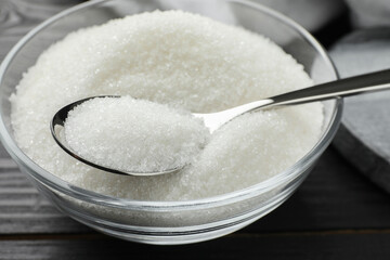 Granulated sugar in bowl and spoon on black wooden table, closeup