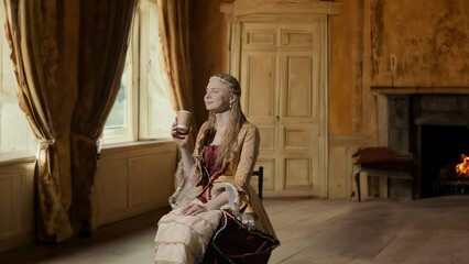 Obraz premium Woman in ancient outfit on background of historic interior. Female in renaissance style dress sitting drinking coffee from paper cup.