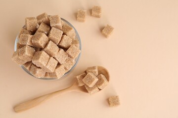 Brown sugar cubes in glass and spoon on beige background, flat lay. Space for text
