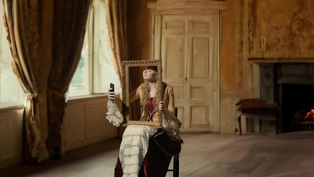 Historical man modern lifestyle advertising. Woman in ancient outfit on background of historic interior. Woman in historical dress holding a framed picture and taking selfies on smartphone.