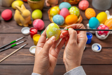 Woman paints Easter eggs at the kitchen wooden table.Happy Easter celebration concept.Colorful...