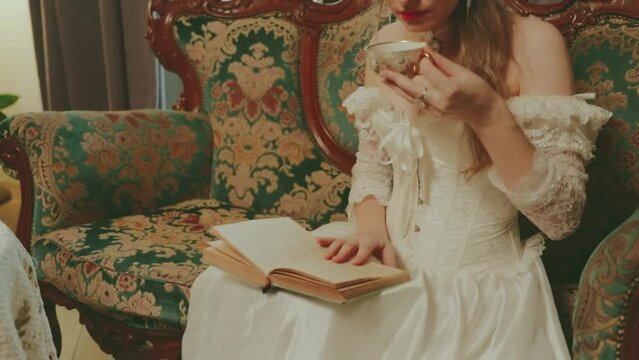 Happy woman drinking hot tea holding cups in hands reading paper novel book. Woman noble lady beauty face lips close-up. retro sexy girl sits on sofa rest royal room white vintage dress old style. 4k