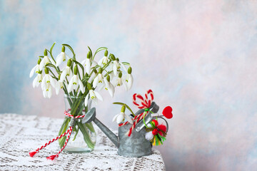 Snowdrop flowers on the table in a vase, watering can, red and white symbol of Martenitsa holiday...