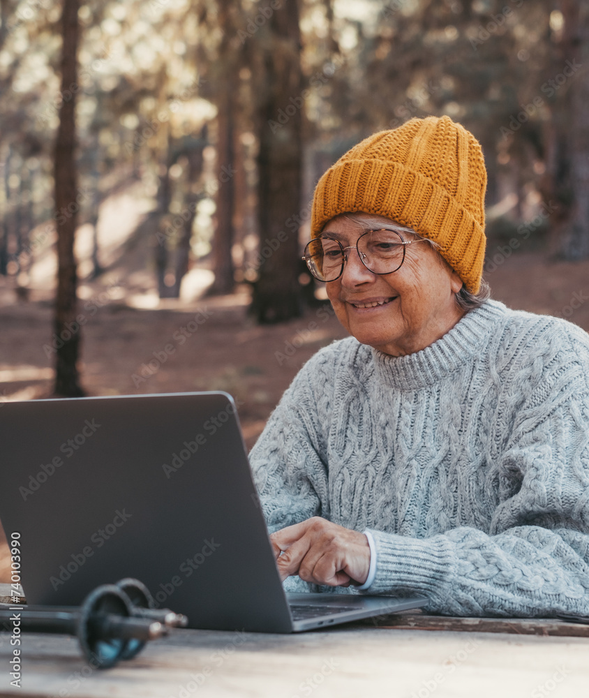 Wall mural Head shot portrait close up of cute one of old middle age person using computer pc outdoors sitting at a wooden table in the forest of mountain in nature with trees around her..