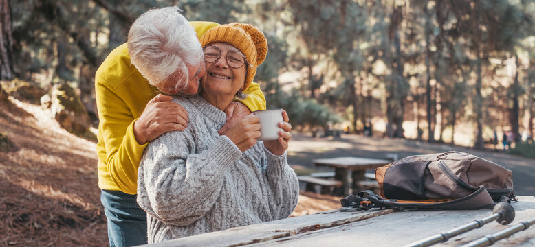 Head Shot Portrait Close Up Of One Middle Age Woman Drinking Coffee Or Tea From A Cup Sitting At Table In The Nature In The Forest Of Mountain With Old Husband Hugging Wife From The Back.