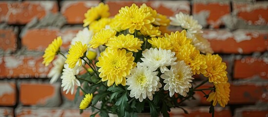 A vase on a table displaying a bunch of yellow and white chrysanthemums with a red brick wall in the background.