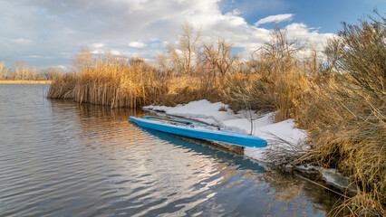 touring stand up paddleboard with a paddle and safety leash on a shore of lake in northern Colorado, winter or early spring scenery with some snow