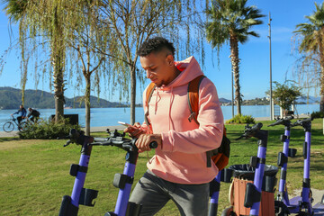 Latin guy in a pink hoodie scanning a QR code to rent an e-scooter in the park. Mixed race young...