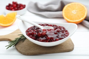 Fresh cranberry sauce in bowl, spoon and rosemary on white wooden table, closeup