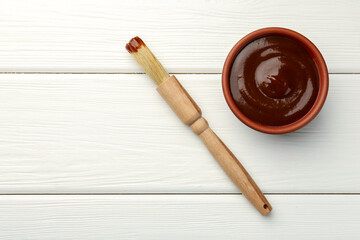 Tasty barbeque sauce in bowl and brush on white wooden table, top view. Space for text