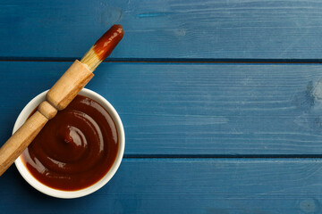 Tasty barbeque sauce in bowl and brush on blue wooden table, top view. Space for text