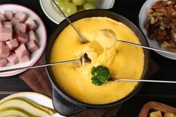 Dipping different products into fondue pot with melted cheese on black wooden table, flat lay