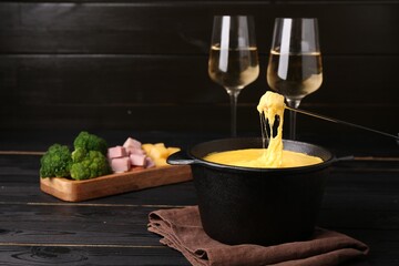 Dipping piece of ham into fondue pot with melted cheese on black wooden table