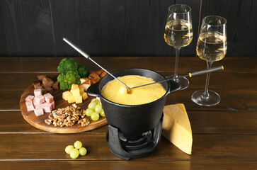 Fondue pot with melted cheese, glasses of wine and different products on wooden table