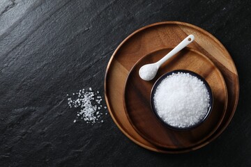 Organic white salt in bowl and spoon on black table, top view. Space for text