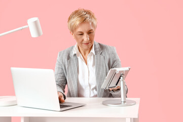 Mature female programmer working with tablet computer and laptop at table on pink background