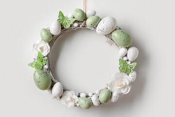 Easter wreath made of flowers, eggs and butterflies with branches hanging on white wall