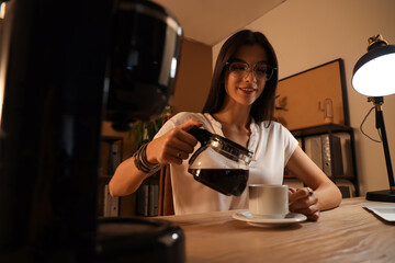 Young businesswoman pouring hot coffee into cup in office at night