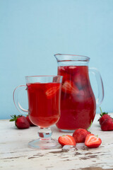 Vertical food photo. Glass tall jug with red compote against blue wall. Transparent cup with strawberry juice on wooden surface of old vintage table. Summer soft drink, lemonade, alcoholic beverage