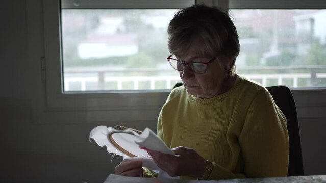 Seventy-year-old slim, blonde Caucasian woman with glasses cross-stitching at home. Looking at drawing and counting stitches and checking colours. ext to a window with natural light. mustard-coloured