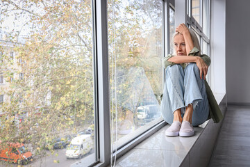 Depressed mature woman sitting near window at home - obrazy, fototapety, plakaty