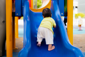 Backside of Asian baby boy climbs the children's slide on playground. Child playing happily in summer or spring times. Toddler playing outdoor. Active kid on colorful slide. Son aged 1 year old.