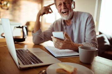 Smiling senior man holding smartphone with laptop on desk at home
