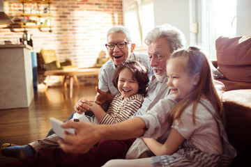 Happy grandparents taking a selfie with grandchildren on the couch at home - obrazy, fototapety, plakaty