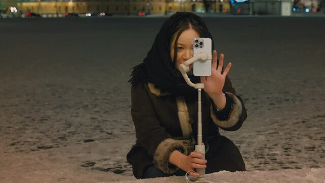 A cute Asian girl takes a video on her phone on a three-axis stabilizer on the night streets of a winter city. A young blogger sets up her mobile phone for an online broadcast. Shooting from a tripod