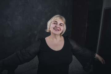 A blonde, short-haired, chubby older woman in black clothes on a solid background of a dark gray concrete.