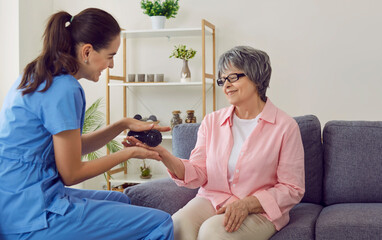Friendly young nurse or physiotherapist holding spiky rubber ball and doing hand massage to happy smiling senior woman who is sitting on sofa in retirement home. Old age, health, physiotherapy concept