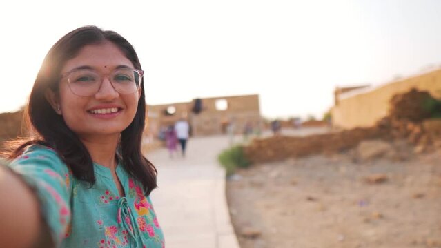 Selfie video of happy Indian young woman smiling at camera at Kuldhara, Rajasthan, India. Positive cheerful girl travel outdoors. Young woman vlogging on vacation. Girl wearing Indian Kurta.