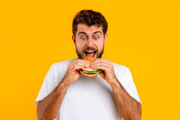 Excited hungry Caucasian man biting delicious cheeseburger against yellow backdrop