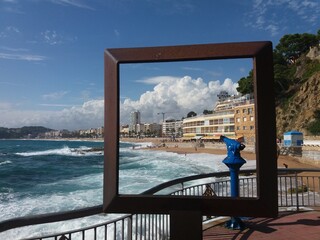 Metal frame. Through it you can see blue tourist binocular, the coast, white clouds and buildings. Around: a sea with white foam, mountains with pine needles, a curved fence and a blue booth