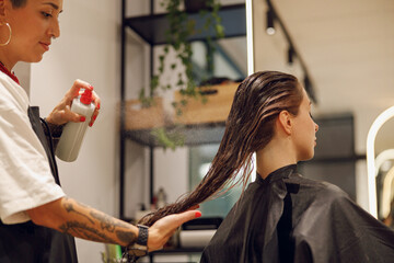 Beautiful hair stylist with tattooed hands applying spray on woman wet hair in beauty salon