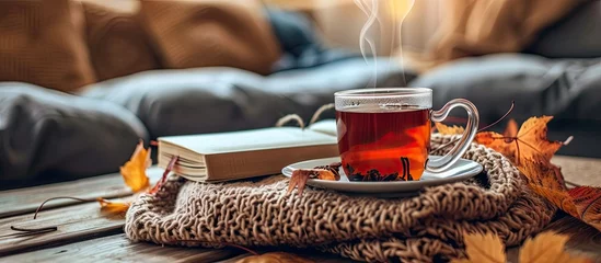 Deurstickers An autumn hygge scene featuring a cup of hot tea and a book resting on a table in a cozy home setting. © AkuAku