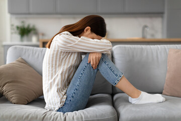 Upset young woman sitting on the sofa with her head down, side view