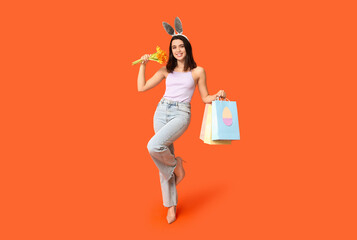 Beautiful young happy woman in bunny ears with shopping bags and bouquet of flowers on orange background. Easter celebration