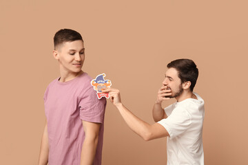 Young man sticking paper clown on his friend's back against brown background. April fool's day celebration