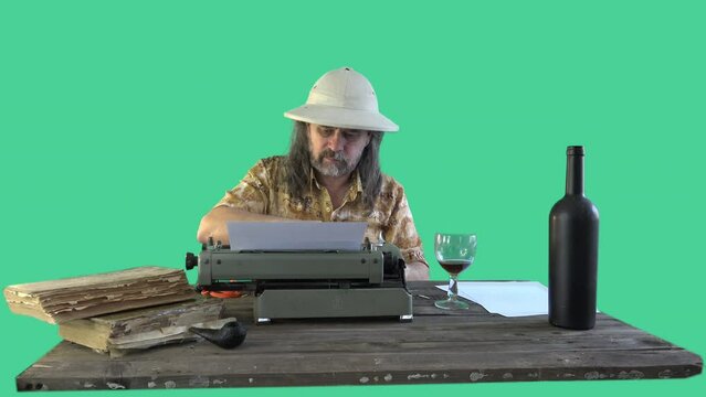 Portrait male traveler (in pith helmet) with long hair writes his memoirs at an old typewriter, contentedly drinks wine from glass at wooden table, there are books and blank sheets of paper on table