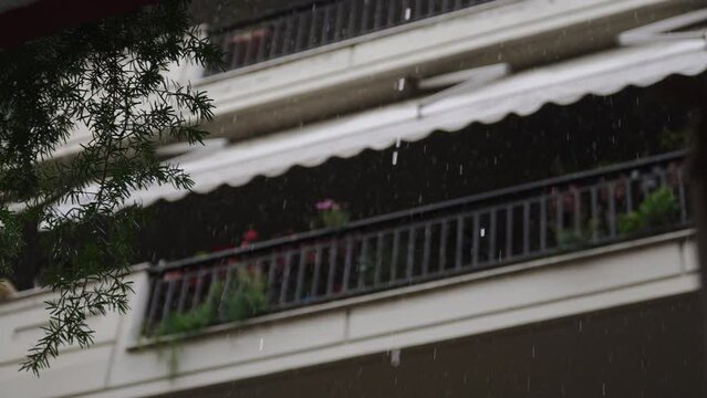 Stream of water flows from overflowing gutter against backdrop of blurred apartment balconies. Rainy day in modern urban area without people. Potted plants grow on balcony. Bad and wet weather.