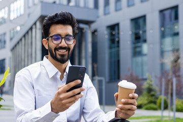 Young man with phone in hands and a cup of hot drink close up, businessman smiling contentedly...
