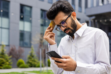 Cheated disappointed sad man outside office wake up, young businessman with phone in hand and bank...