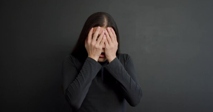 Deeply shocked confused young Asian man in black sweater looking at camera with terrified expression and closing mouth, face, hiding behind long dark hair Studio shot isolated background. fear, fobia