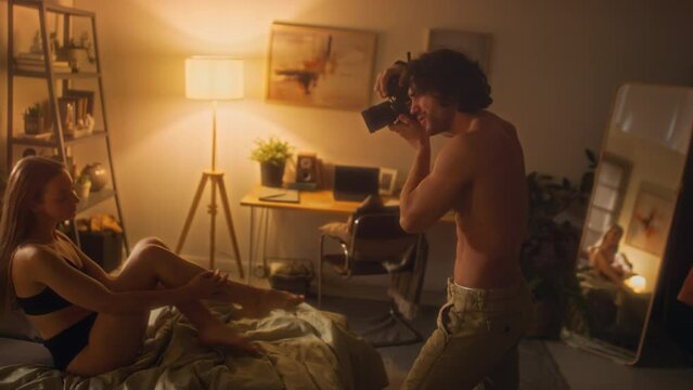 Young shirtless man taking pictures of girlfriend in underwear posing on bed in cozy room with warm light