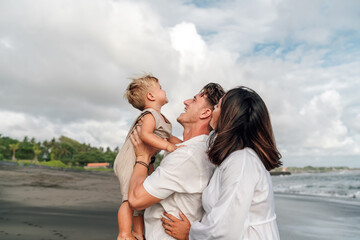 Family embrace on the beach: Father holding a laughing toddler aloft, mother looking at them lovingly, against a backdrop of sea and sky, capturing a moment of joy and parental love